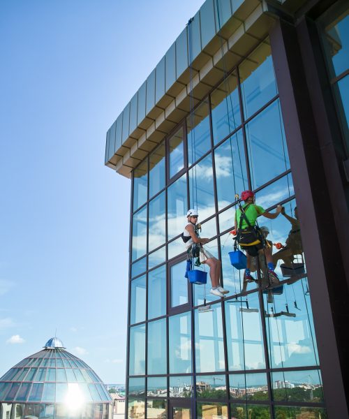 Industrial mountaineering. Male washers hanging on ropes and using cleaning tools while washing building windows. Two men window cleaners in safety helmets working together at high-rise building.
