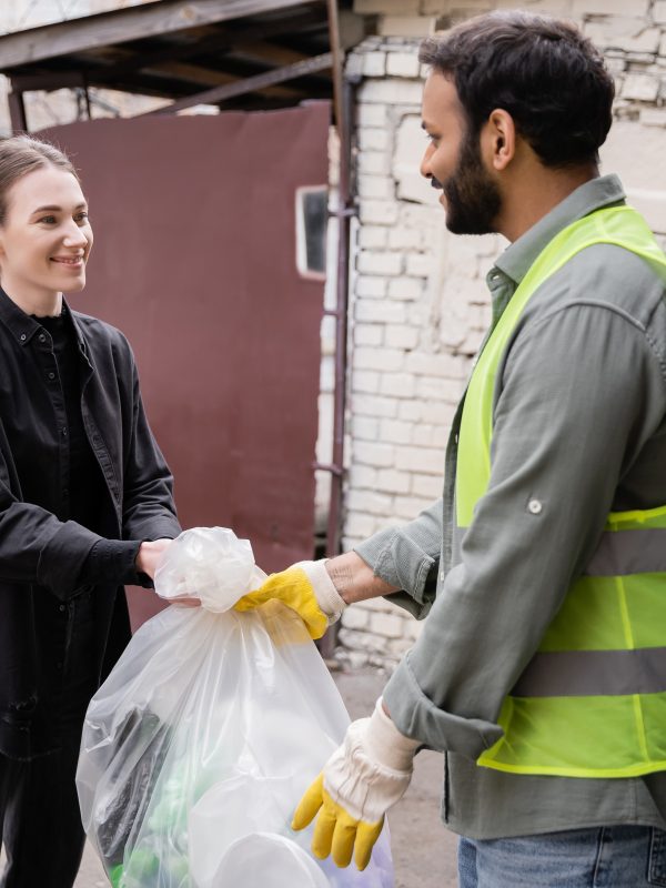 Smiling young volunteer giving plastic bag with trash to indian worker in protective vest and gloves outdoors in waste disposal station, garbage sorting and recycling concept